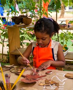 Menina de avental pintando pedaços de papelão no ateliê ao ar livre
