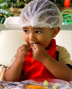 Menino de touca e avental provando uma fruta na aula de culinária