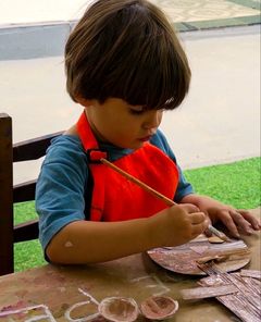 Menino de avental pintando uma peça de papelão com pincel