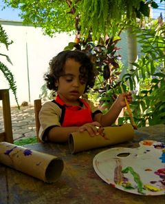 Menino de avental pintando um rolo de papelão no ateliê ao ar livre